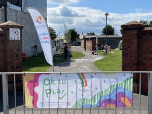 A colourful banner with words "ok to play" hung on railings in front of apark.