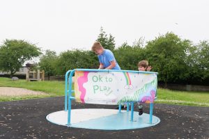 In a playpark, on a roundabout hangs a banners with the words of to play. A young boy is being pushed on the roundabout by a man, both are smiling.