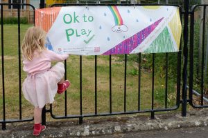 A banner on railings reads Ok to Play. A young girl plays at climbing the railings next to it.