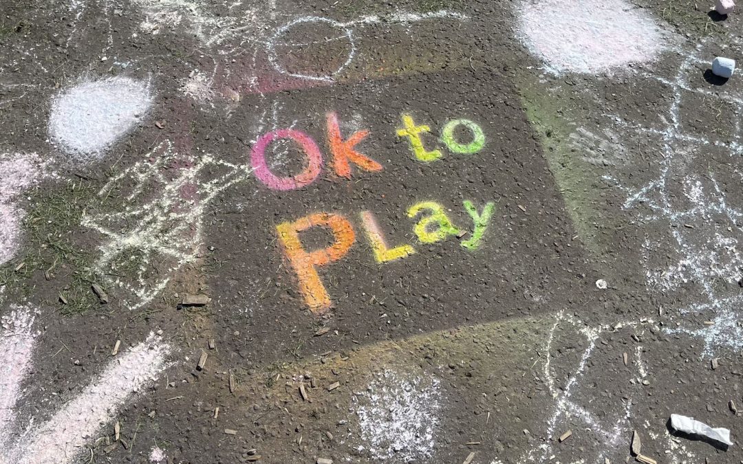the words Ok to Play are stenciled in rainbow colours on a pavement.