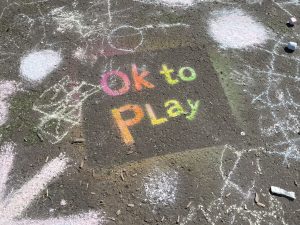 the words Ok to Play are stenciled in rainbow colours on a pavement.