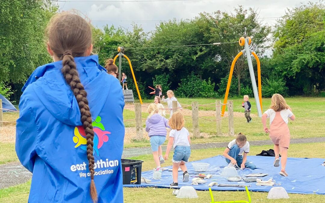 To the left a Play Ranger stands with her back to us, wearing a blue jacket with the East Lothian Play Association logo. In the background is a playpark with children playing and a blue tarpaulin on the ground for water play.